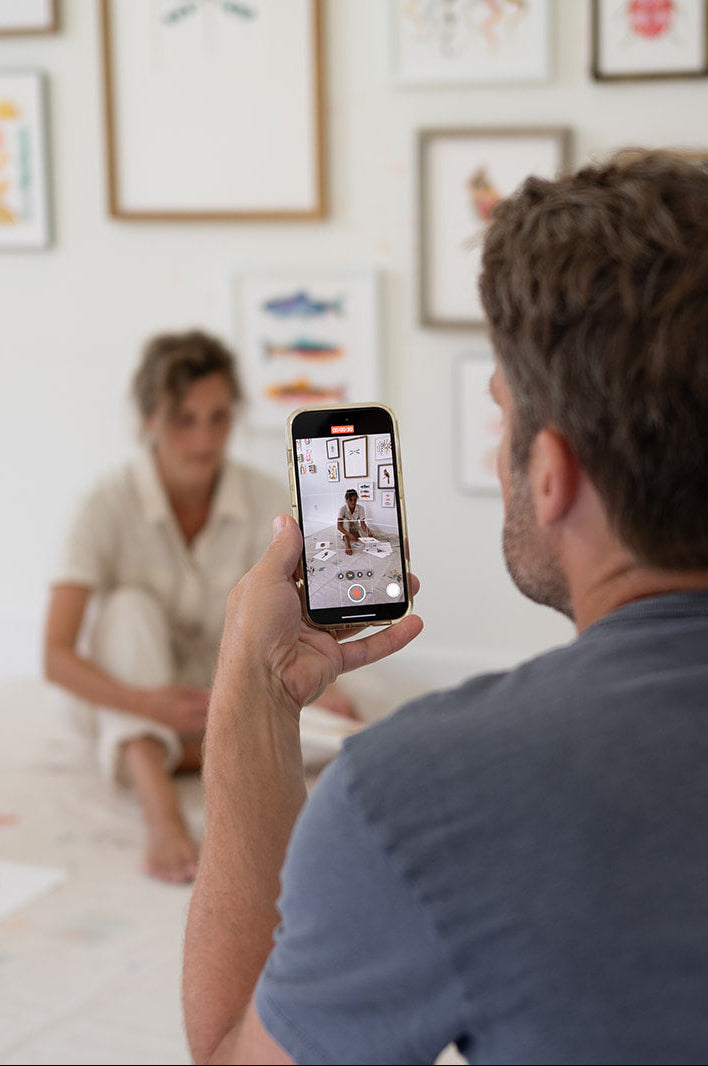 Man taking a photo of a woman with a smartphone in a room with framed pictures on the wall.